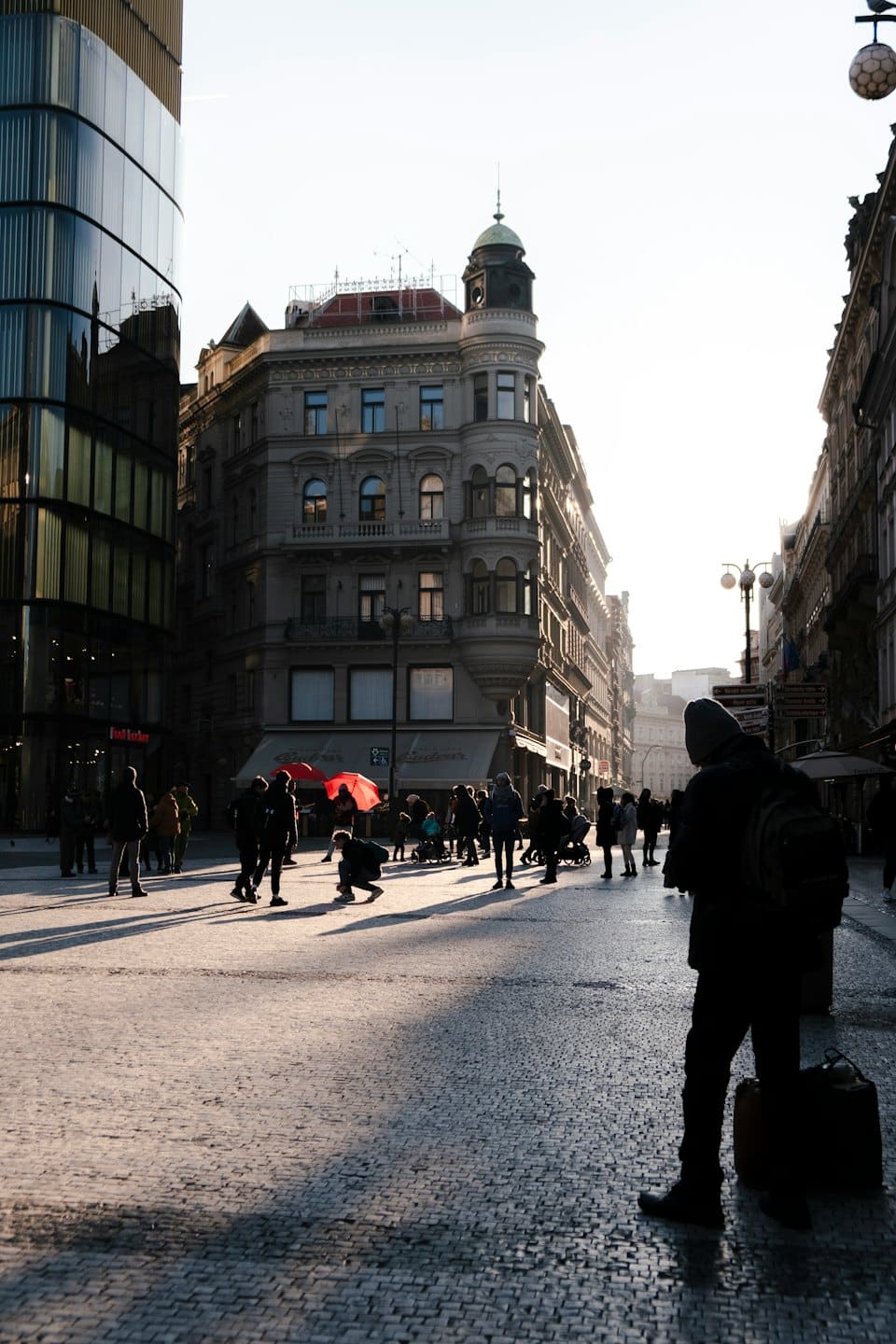 Pedestrians walk along a cobblestone street between modern and historic buildings.