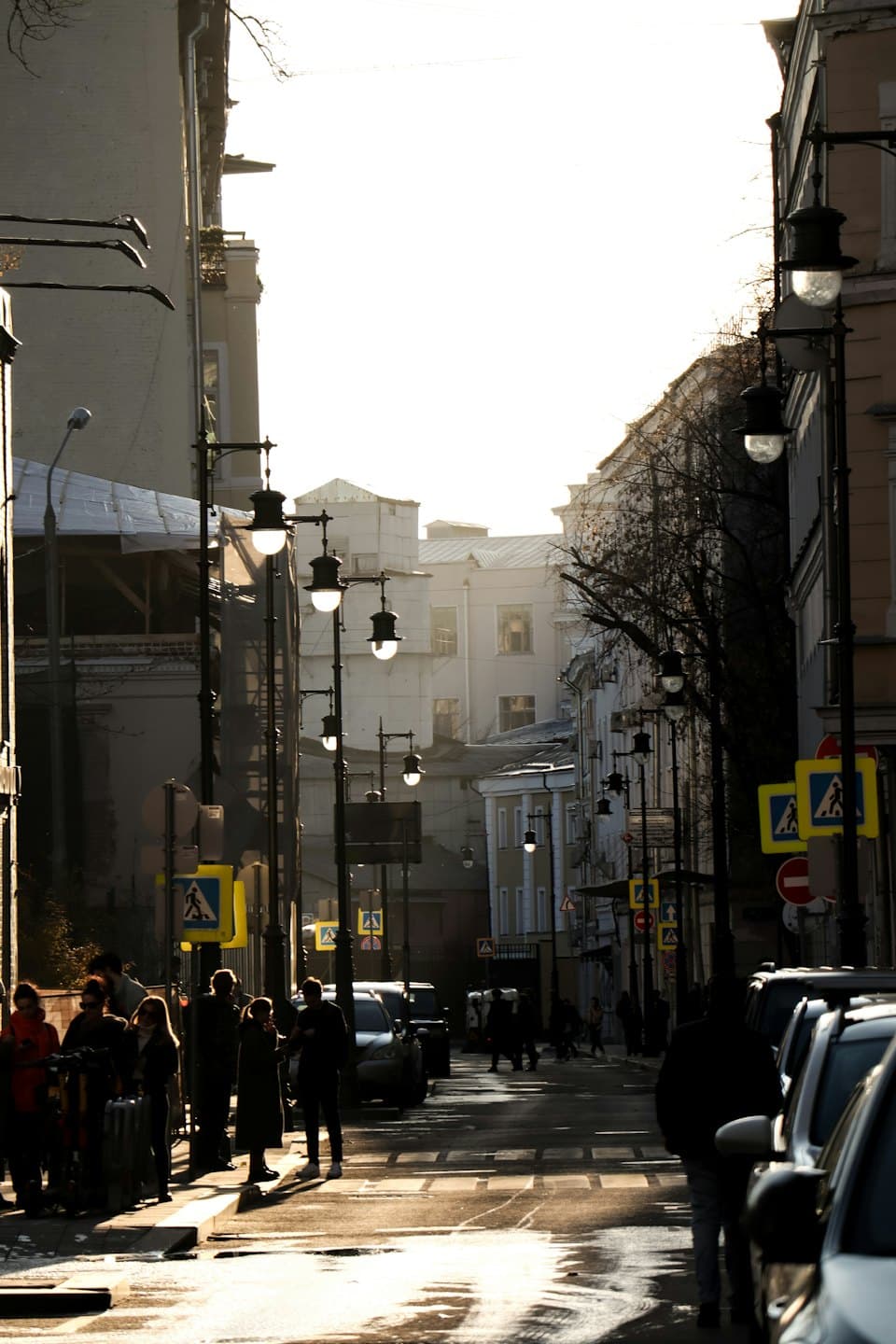 Pedestrians walk along a wet city street between tall buildings.
